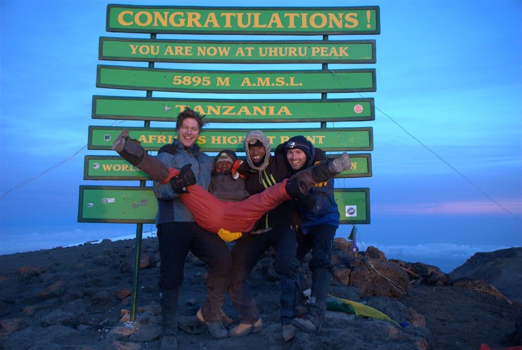 A group of climbers celebrates in front of the Uhuru Peak summit sign on Mount Kilimanjaro.