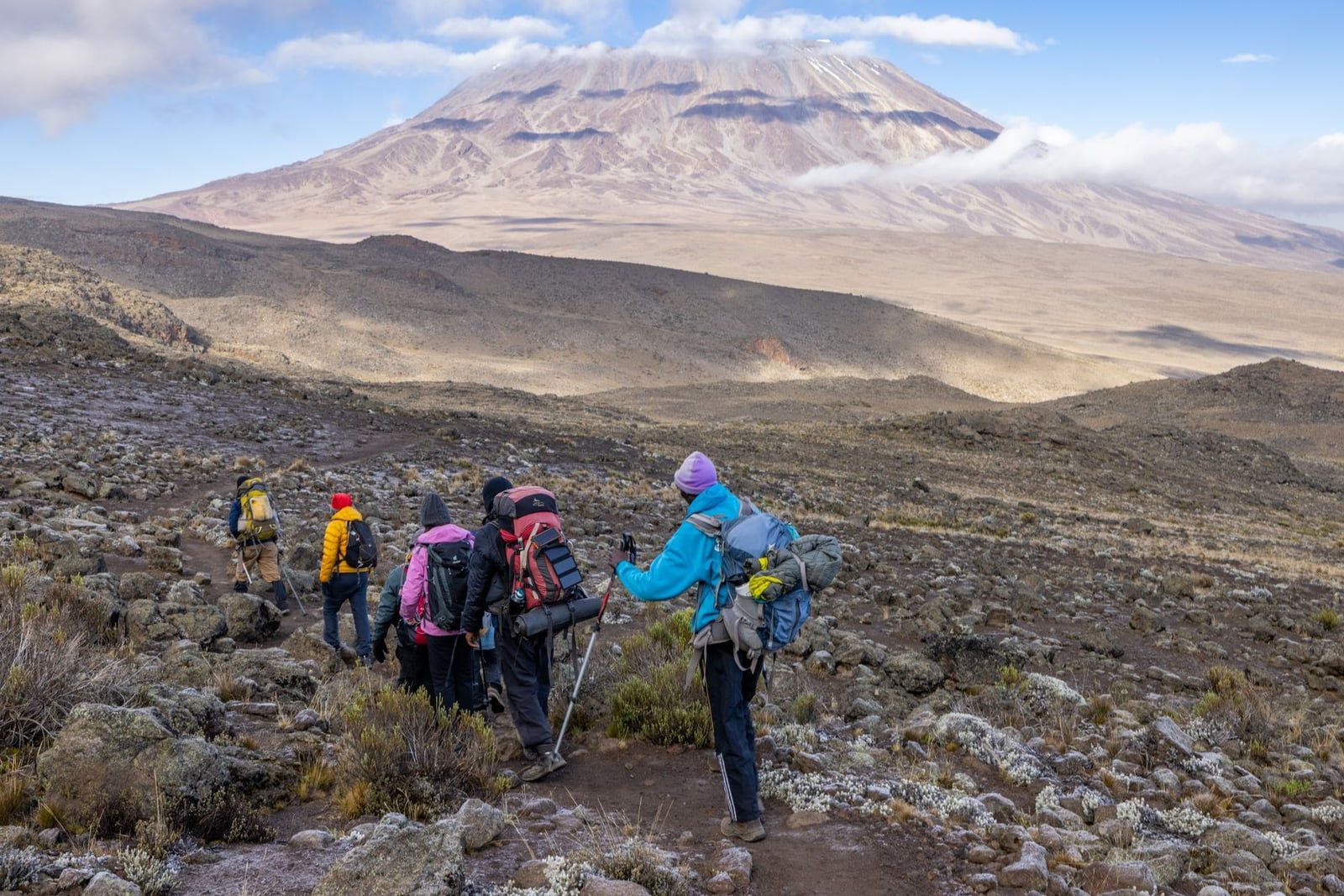 Trekkers carrying backpacks hike along a rocky mountain trail on Mount Kilimanjaro with the volcanic peak in the distance.