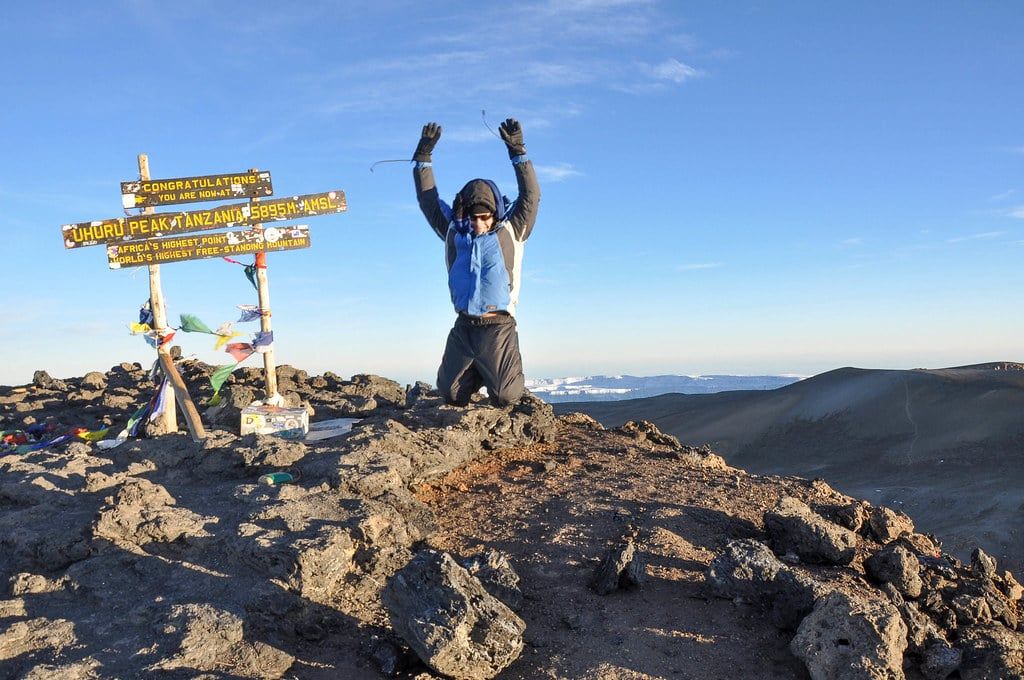 A hiker jumps for joy beside the wooden summit signpost at Uhuru Peak on Mount Kilimanjaro.