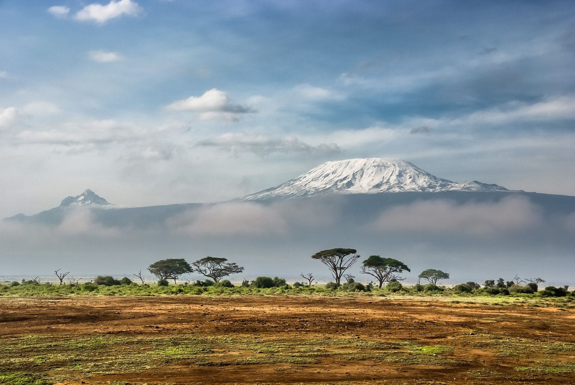Snow-capped Mount Kilimanjaro and its rugged Mawenzi peak rise above a band of clouds and the African savanna.